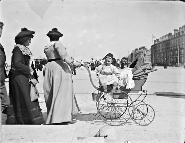 Two women and one man walking with perambulator and infant. Women pushing perambulator has face turned away from the camera, infant sitting&nbsp;...