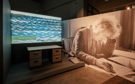 Seamus Heaney's writing desk with video above it. To the right there is a large black and white photograph of Seamus Heaney at a desk, reading.