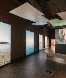 Section of 'Seamus Heaney: Listen Now Again' exhibition space. To the left is a row of scenic images of bodies of water. To the right a visitor sits on a bench watching a video of Seamus Heaney speaking.