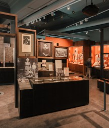 Section of 'Seamus Heaney: Listen Now Again' exhibition. A low brown case with black and white images of Seamus Heaney taken during his childhood and early adult years above it. He is shown graduating from college, with primary school classmates and with a family member. A visitor is shown stooped over looking at a different case in the background.