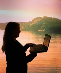 The profile of a woman is seen using a laptop against a picturesque background of a lake with land in the distance 