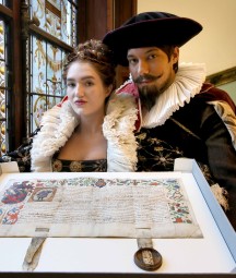 Two actors in period costume pose with grant of arms next to stained glass window