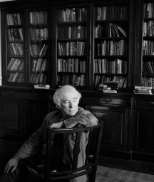 Black and white photograph of Seamus Heaney sitting in a chair with bookshelves behind him