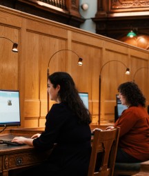 Suíonn triúr ban ag déanamh taighde ar scáileáin ríomhaire taobh istigh de phríomhsheomra léitheoireachta an NLI