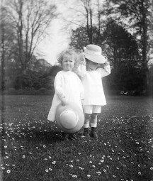 Two toddlers with bonnets standing in field surrounded by daisies