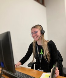 Photograph of blonde-haired young woman looking at computer with headset on. 