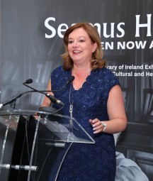 Photograph of woman with brown hair wearing blue dress speaking into microphone. 