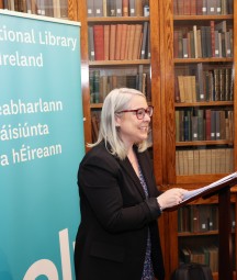 Woman speaking from a podium with bookshelves in the background