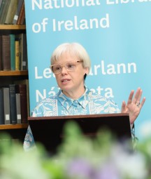 A woman giving a speech in front on a blue banner and bookshelves