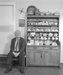 Seamus Heaney sitting in a wooden chair in a rural kitchen next to a cabinet filled with crockery