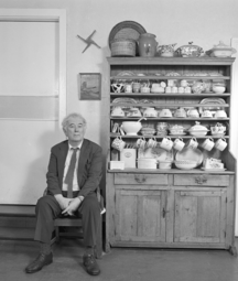Seamus Heaney sitting in a wooden chair in a rural kitchen next to a cabinet filled with crockery