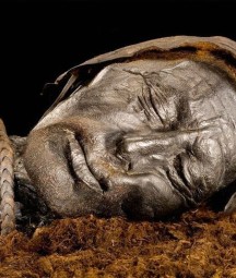 Close-up photograph of the preserved head of the Tollund Man, a naturally mummified body found in a Danish peat bog. His eyes are closed and his face appears peaceful, with detailed features and leathery skin preserved by the bog’s conditions. A rope noose is visible around his neck, and he wears a pointed leather cap.