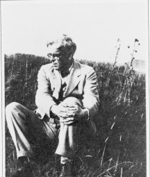 Black and white photograph of poet W. B. Yeats sitting in a grassy field. He is dressed in a light-colored suit and glasses, with his arms wrapped around one knee, gazing to his left. The setting is natural and serene, with tall grass and wild plants in the background.