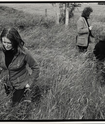 Black-and-white photograph of a young Marie Heaney walking through tall grass in a rural field, looking down as wind bends the grass around her. In the background, Seamus Heaney stands near a fence with two young children, all partially obscured by the grass, with open farmland stretching behind them.
