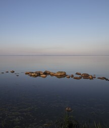 Still water at Lough Neagh with a line of smooth rocks breaking the surface, reflected clearly in the calm lake. The horizon is faint in the distance beneath a wide, pale blue sky, creating a quiet, minimalist scene.
