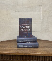 A stack of three copies of The Poems of Seamus Heaney sits on a wooden table against a pale stone wall. Two copies are laid flat with one standing upright on top, showing the book’s dark, textured cover design and title.
