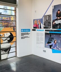 Music exhibition interior featuring large concert photos, including a singer on a window graphic and a wall display about Live Aid at Wembley, with text panels and a QR code.