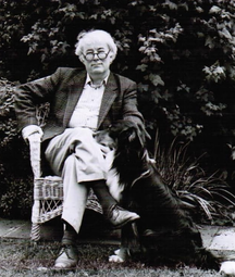 A black-and-white photo of Seamus Heaney sitting in a wicker chair in a garden. He wears a jacket, collared shirt, and trousers, sitting with one leg crossed over the other. Carlo the dog sits beside him, looking up at him. Dense leafy bushes fill the background, and the ground is grassy.