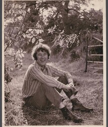 Black-and-white photograph of a young Seamus Heaney sitting on grass in a rural field, smiling at the camera. He wears a striped long-sleeve shirt, trousers, and lace-up boots, with his arms loosely wrapped around his knees. Tree branches with leaves hang above him, and a wooden gate stands in the background. 