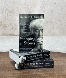 A stack of three books titled Stepping Stones: Interviews with Seamus Heaney by Dennis O’Driscoll sits on a wooden table against a textured, off-white wall, with one copy standing upright on top displaying a black-and-white portrait on the cover.