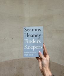 Hand holding a light blue copy of Finders Keepers by Seamus Heaney against a curved stone wall background.