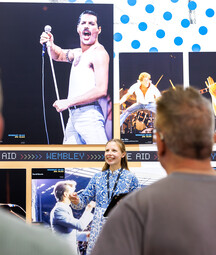 A female tour guide smiles and gestures while speaking to a small group of visitors in front of a large exhibition wall featuring iconic concert photographs. A prominent image of Freddie Mercury, shirtless and performing with a microphone dominates the display, with additional photos of other musicians arranged around it. The visitors are seen from behind, focused on the guide during the tour.