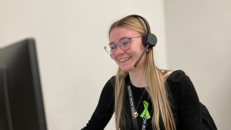 Photograph of blonde-haired young woman looking at computer with headset on. 