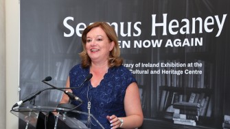 Photograph of woman with brown hair wearing blue dress speaking into microphone.