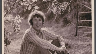 Black and white photograph of man, Seamus Heaney, sitting on grass in a striped shirt. 