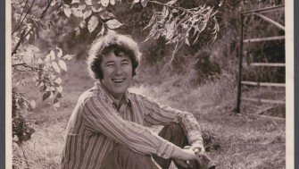 Black and white photograph of man, Seamus Heaney, sitting on grass in a striped shirt.