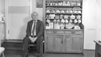 Seamus Heaney sitting in a wooden chair in a rural kitchen next to a cabinet filled with crockery