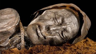 Close-up photograph of the preserved head of the Tollund Man, a naturally mummified body found in a Danish peat bog. His eyes are closed and his face appears peaceful, with detailed features and leathery skin preserved by the bog’s conditions. A rope noose is visible around his neck, and he wears a pointed leather cap.