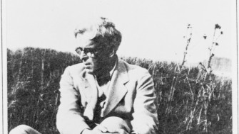 Black and white photograph of poet W. B. Yeats sitting in a grassy field. He is dressed in a light-colored suit and glasses, with his arms wrapped around one knee, gazing to his left. The setting is natural and serene, with tall grass and wild plants in the background.