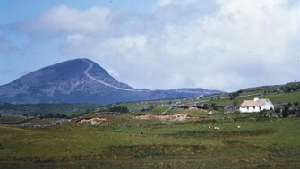 A scenic landscape featuring a large, rounded mountain with a visible trail leading to its summit, surrounded by green fields and stone walls. In the foreground, a white cottage with a thatched roof sits among rolling grassy hills under a partly cloudy sky.