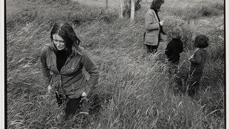 Black-and-white photograph of a young Marie Heaney walking through tall grass in a rural field, looking down as wind bends the grass around her. In the background, Seamus Heaney stands near a fence with two young children, all partially obscured by the grass, with open farmland stretching behind them.