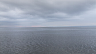 A wide view of the Flaggy Shore stretching to the horizon beneath a cloudy sky, with rounded grey stones in the foreground at the water’s edge.”