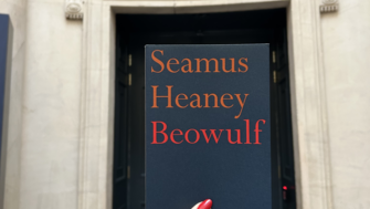 A hand holding up a dark blue copy of Beowulf against a marble doorway in the background
