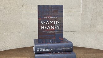 A stack of three copies of The Poems of Seamus Heaney sits on a wooden table against a pale stone wall. Two copies are laid flat with one standing upright on top, showing the book’s dark, textured cover design and title.