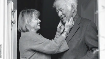 Black-and-white photo of Marie Heaney standing in a doorway, smiling warmly as she gently holds Seamus Heaney's face. Heaney, wearing a suit jacket, smiles back with one hand in his pocket, sharing an affectionate, intimate moment.