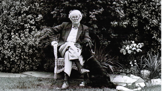 A black-and-white photo of Seamus Heaney sitting in a wicker chair in a garden. He wears a jacket, collared shirt, and trousers, sitting with one leg crossed over the other. Carlo the dog sits beside him, looking up at him. Dense leafy bushes fill the background, and the ground is grassy.