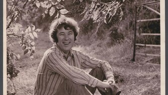 Black-and-white photograph of a young Seamus Heaney sitting on grass in a rural field, smiling at the camera. He wears a striped long-sleeve shirt, trousers, and lace-up boots, with his arms loosely wrapped around his knees. Tree branches with leaves hang above him, and a wooden gate stands in the background. 