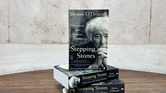 A stack of three books titled Stepping Stones: Interviews with Seamus Heaney by Dennis O’Driscoll sits on a wooden table against a textured, off-white wall, with one copy standing upright on top displaying a black-and-white portrait on the cover.