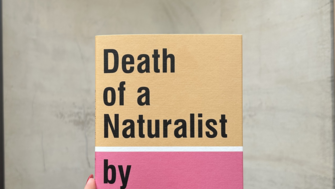 A hand with red-painted nails holds a book titled “Death of a Naturalist by Seamus Heaney.” The cover is split into two colors—beige on top and pink on the bottom—with bold black text. The background is a plain, light gray wall with a curved wooden surface at the bottom.