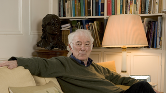 Seamus Heaney sitting on a yellow sofa in his home in front of a bookcase