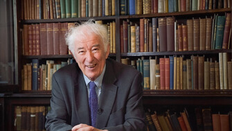 Seamus Heaney sitting smiling in front of a bookcase of old books