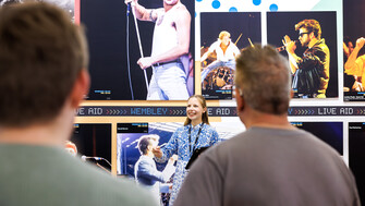 A female tour guide smiles and gestures while speaking to a small group of visitors in front of a large exhibition wall featuring iconic concert photographs. A prominent image of Freddie Mercury, shirtless and performing with a microphone dominates the display, with additional photos of other musicians arranged around it. The visitors are seen from behind, focused on the guide during the tour.