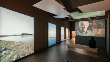 Section of 'Seamus Heaney: Listen Now Again' exhibition space. To the left is a row of scenic images of bodies of water. To the right a visitor sits on a bench watching a video of Seamus Heaney speaking.