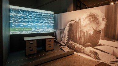Seamus Heaney's writing desk with video above it. To the right there is a large black and white photograph of Seamus Heaney at a desk, reading.
