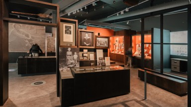 Section of 'Seamus Heaney: Listen Now Again' exhibition. A low brown case with black and white images of Seamus Heaney taken during his childhood and early adult years above it. He is shown graduating from college, with primary school classmates and with a family member. A visitor is shown stooped over looking at a different case in the background.