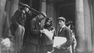 A group of men stands on the steps of Harcourt Street station, where two share a newspaper and young man holds a stack of newspapers for sale under his arm 